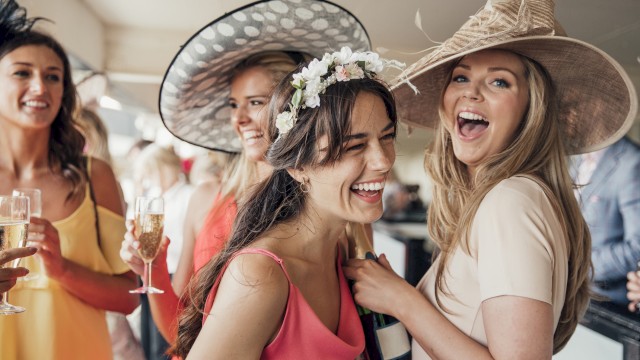 A group of women in stylish hats and dresses enjoy a joyful moment together, while holding glasses of champagne at a social event.