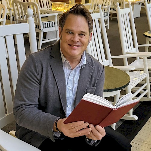 A person is sitting in a white rocking chair, holding an open red book, in an area with several chairs and large windows.