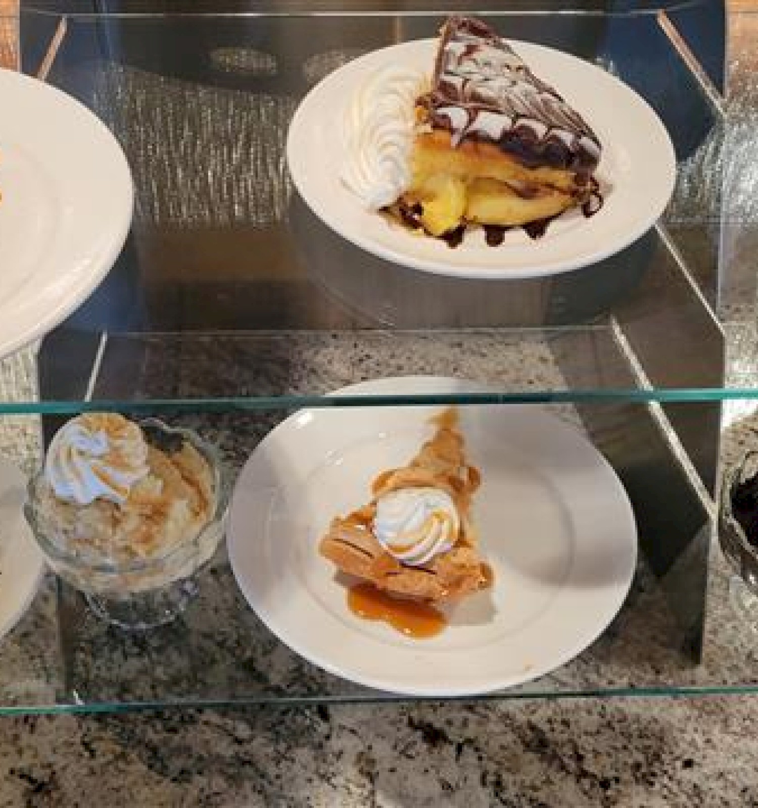 A glass case of assorted desserts: lemon bars, chocolate-topped tarts, peach pastries, whipped cream-topped slices, and a few pies, plus oranges and cakes.