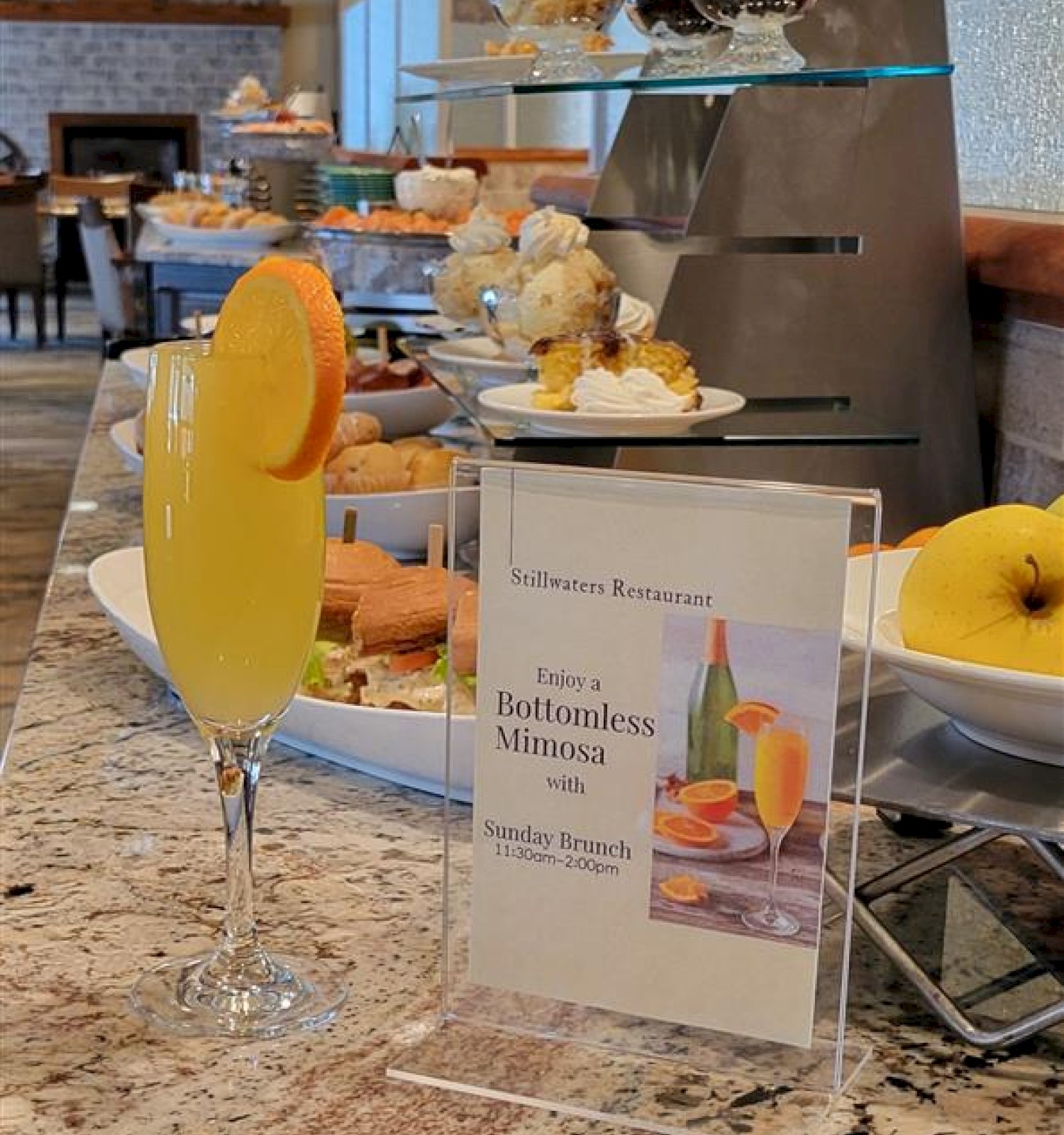 A dessert café counter with pastries, a glass of orange juice garnish, and a menu card for “Gluten-Free Benedict” or similar.