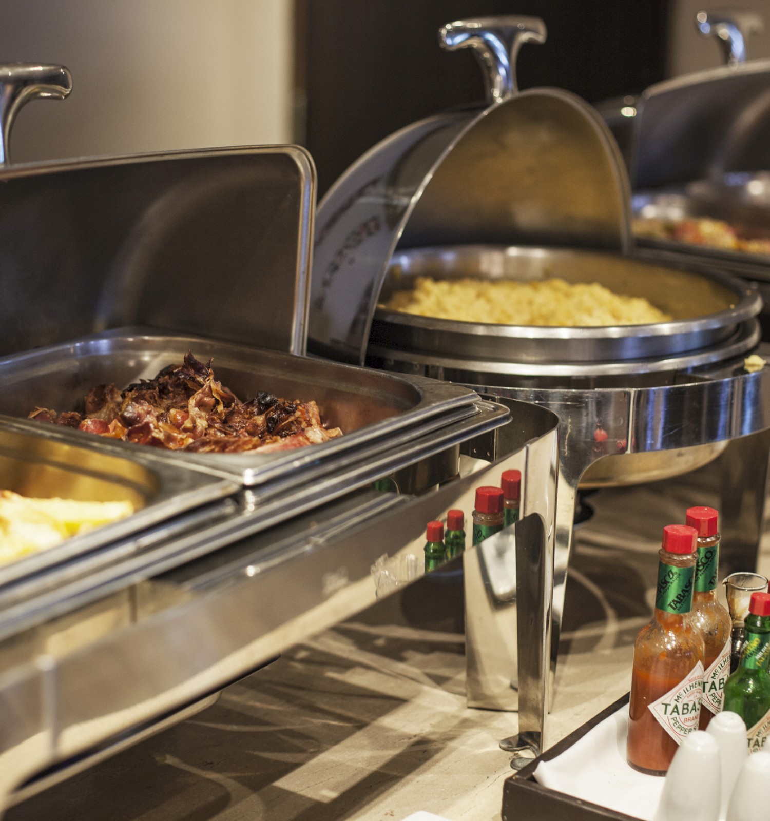 The image shows a buffet setup with trays of food, including pasta and meats, alongside condiments like Tabasco and salt shakers.