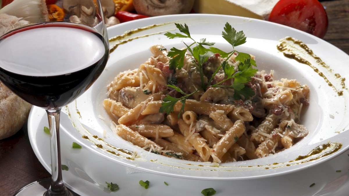 A plate of creamy pasta garnished with parsley, surrounded by ingredients, paired with a glass of red wine.