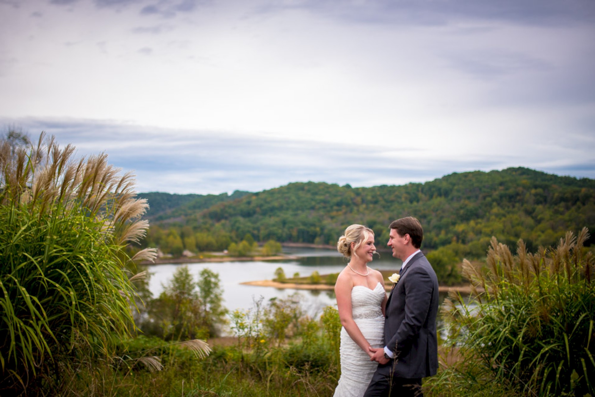 A couple stands outdoors, dressed formally, with a scenic background of a lake and hills, surrounded by greenery.