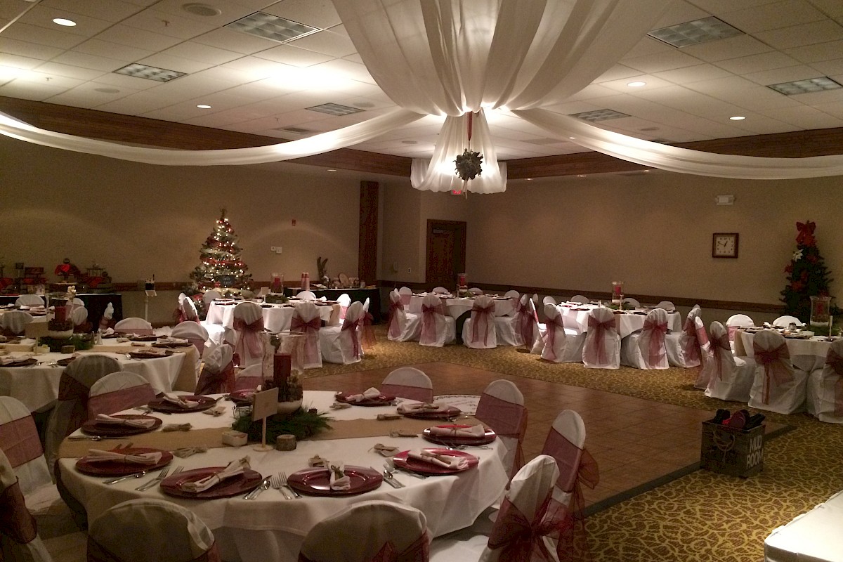 A banquet hall decorated for an event, featuring elegantly set round tables, red accents, and Christmas trees in the background.