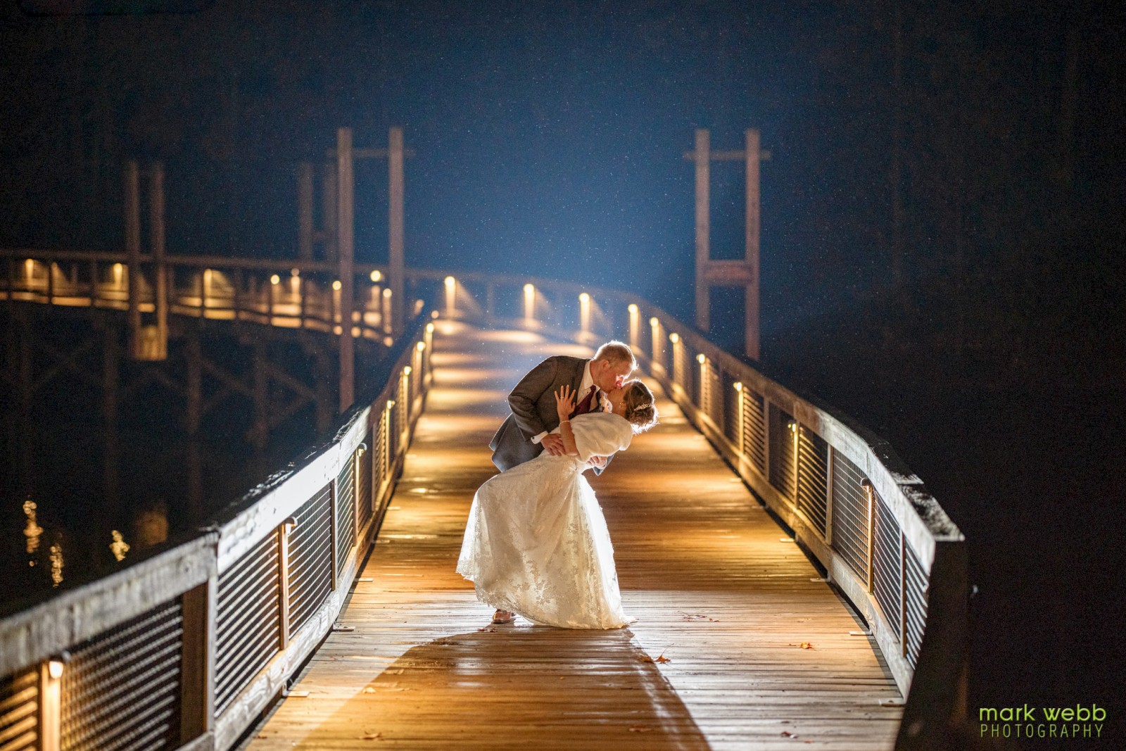 A couple is romantically posing on a lit boardwalk at night, with one partner dipping the other under a night sky.
