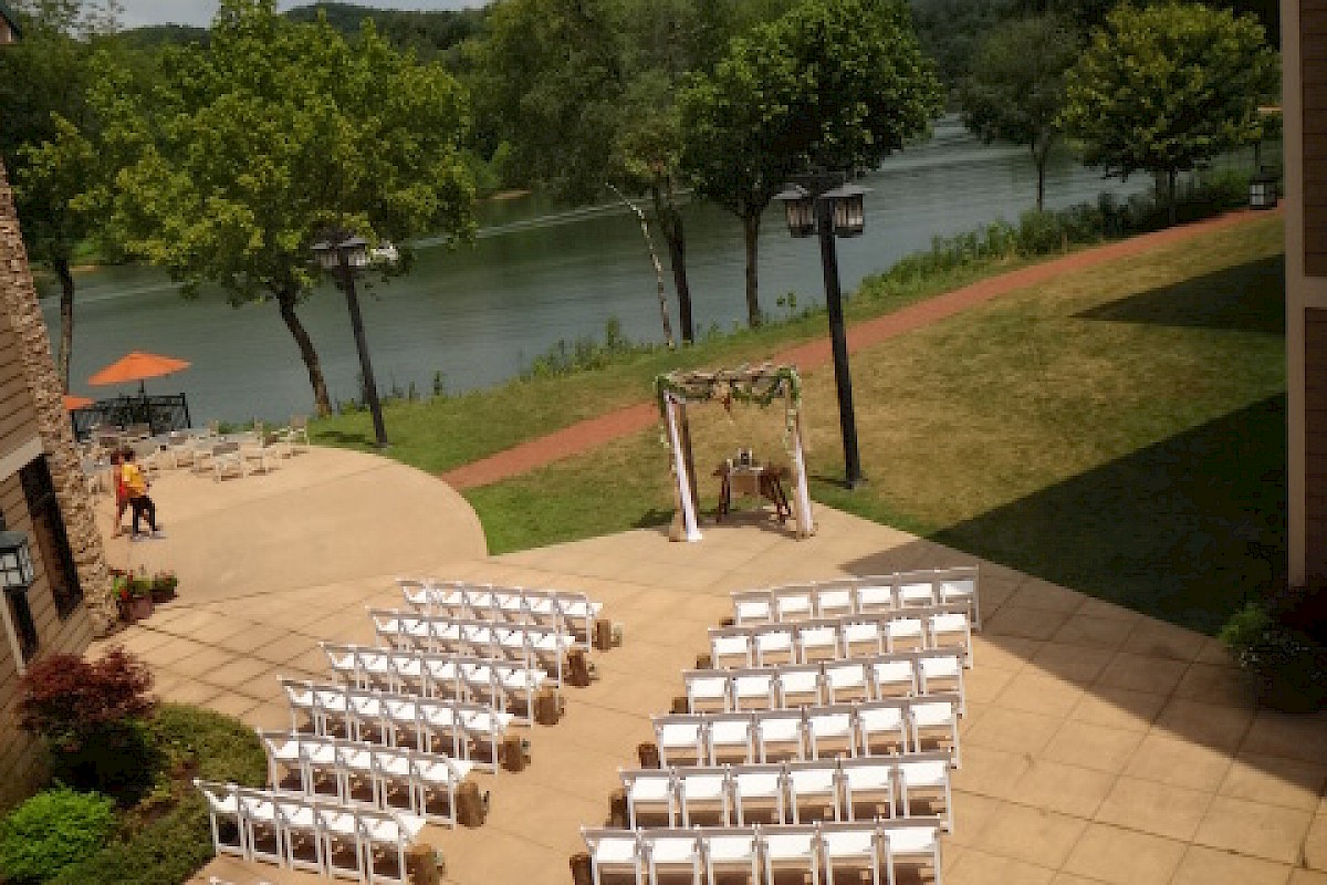 An outdoor wedding setup with white chairs facing an arch near a lake, surrounded by greenery, on a sunny day.