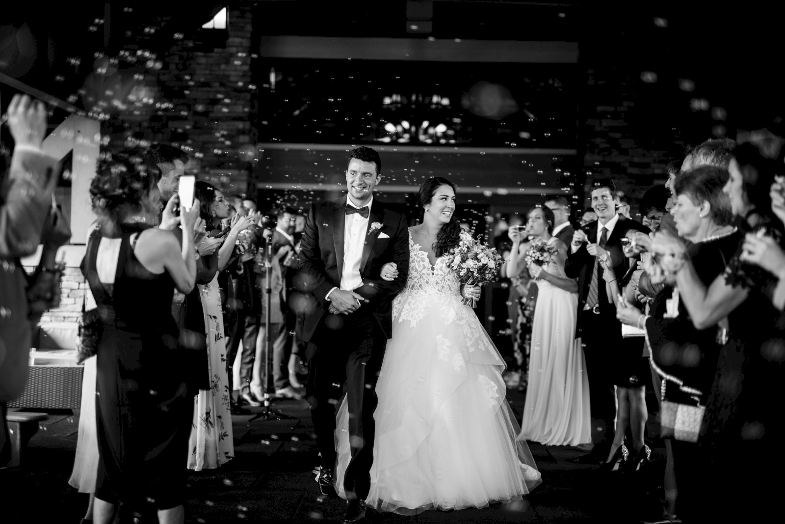 A bride and groom are walking through a crowd of cheering guests. They are dressed formally, and everyone seems to be celebrating.