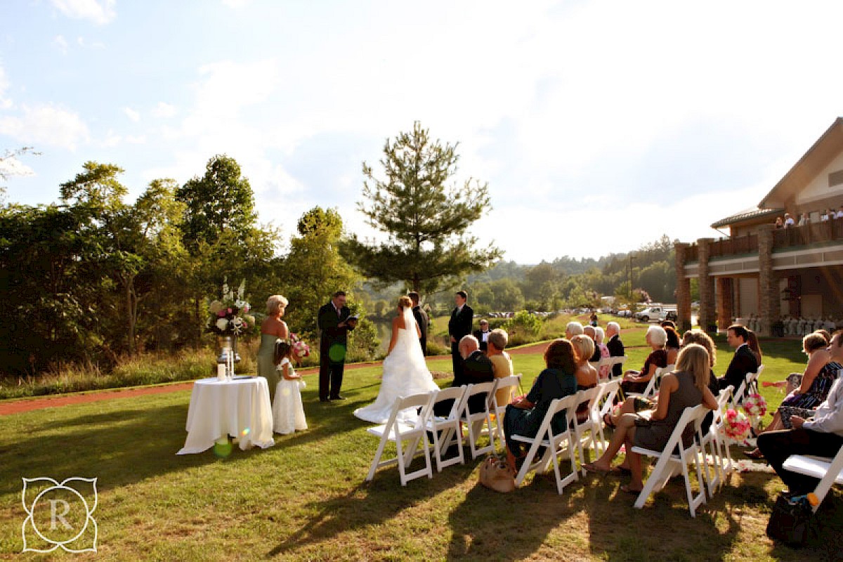 A wedding ceremony is taking place outdoors on a sunny day, with guests seated on white chairs and the couple standing at the altar.