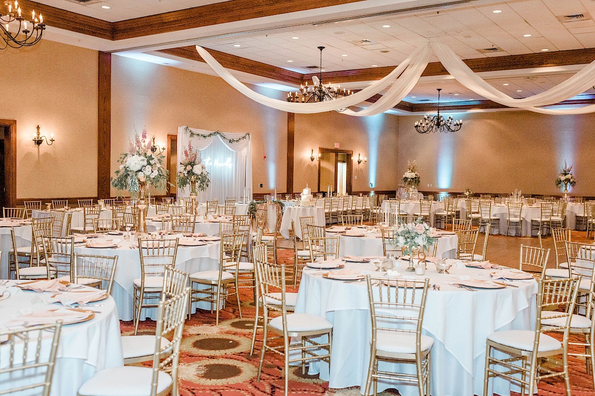 A decorated banquet hall with round tables, white tablecloths, golden chairs, floral centerpieces, and draped fabric on the ceiling.