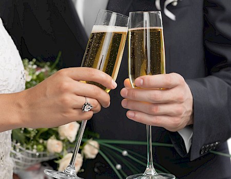 A couple in formal attire toasting with champagne flutes, bouquet of flowers visible, suggesting wedding celebration.