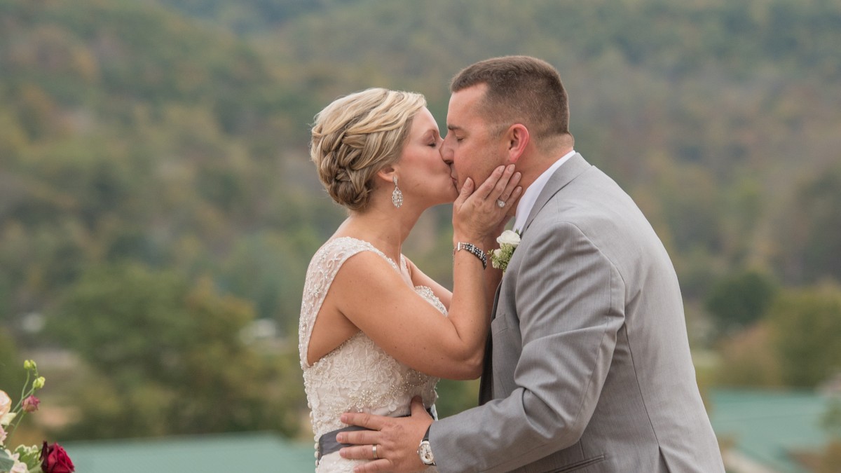 A bride and groom share a kiss outdoors with a scenic mountain backdrop, wearing wedding attire and displaying affection.