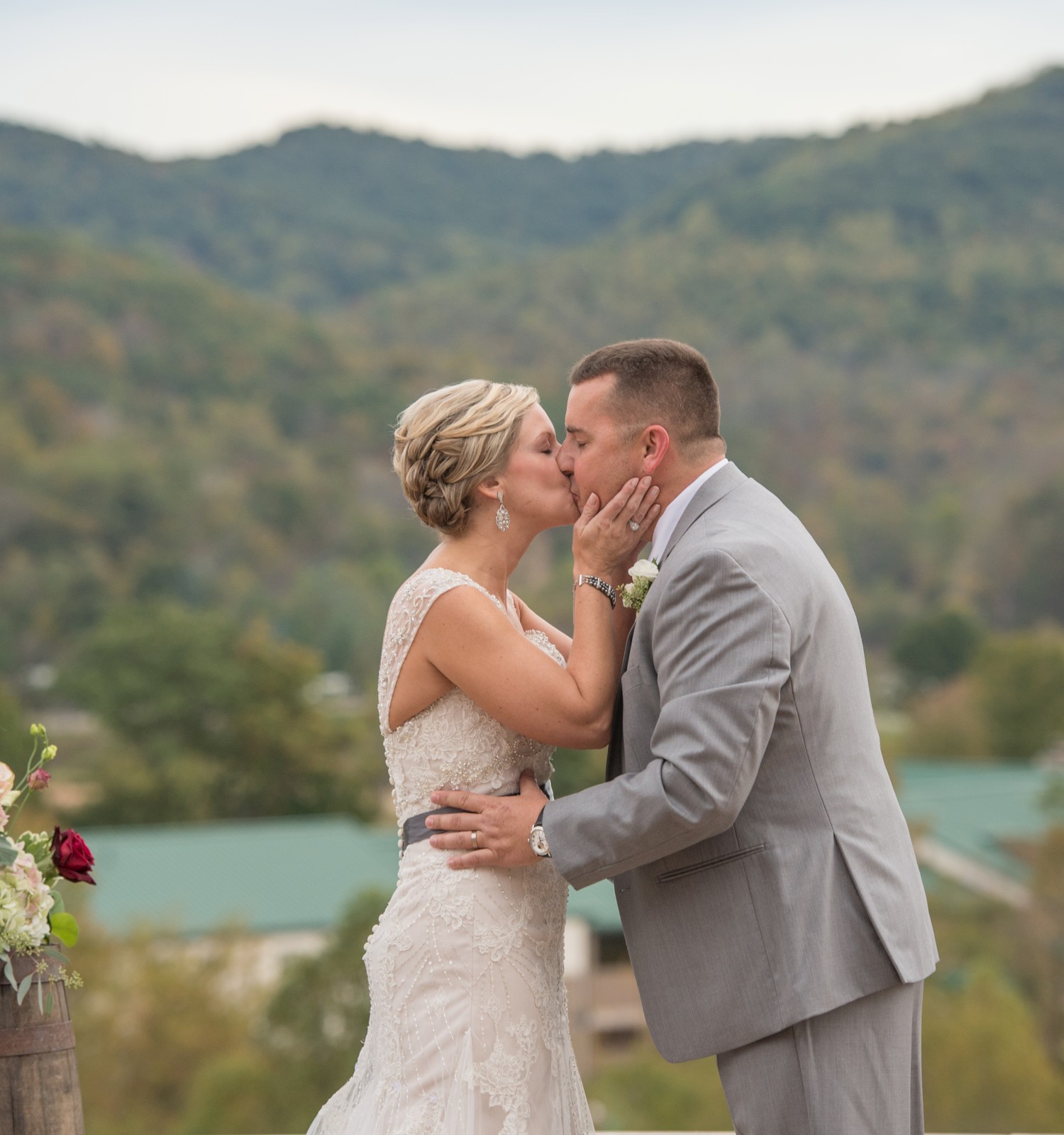 A bride and groom share a kiss outdoors with a scenic mountain backdrop, wearing wedding attire and displaying affection.