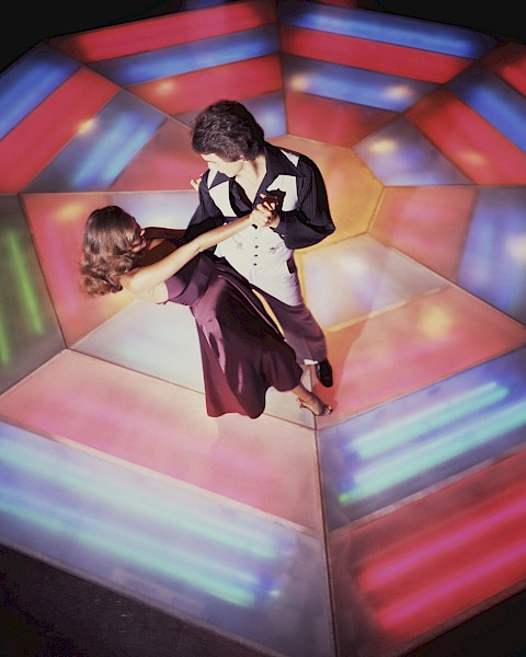 A couple is dancing on a colorful, illuminated floor, with patterns in vibrant hues, creating a lively and nostalgic disco atmosphere.