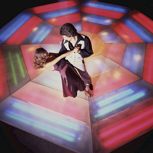 A couple is dancing on a colorful, illuminated floor, with patterns in vibrant hues, creating a lively and nostalgic disco atmosphere.