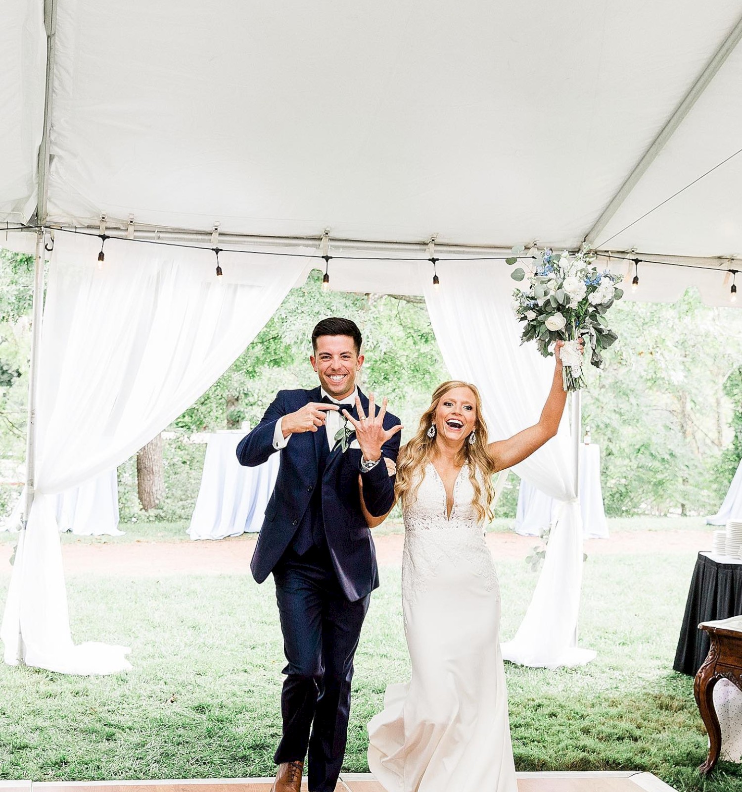 A couple is joyfully entering a tented venue; the woman holds a bouquet and both are smiling, likely celebrating a special occasion.