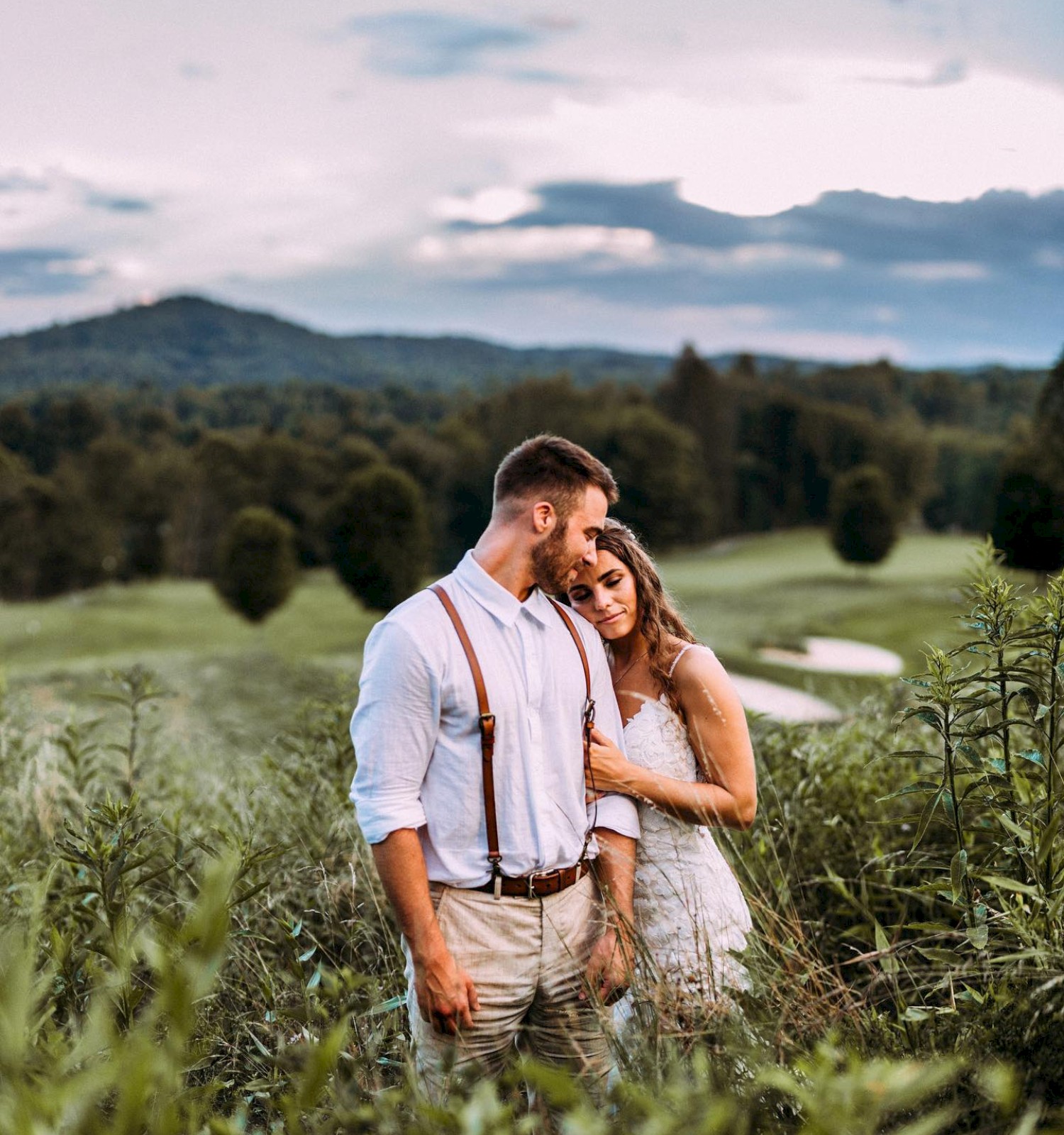 A couple stands in a grassy field, embracing amidst lush greenery with a scenic, hilly background under a cloudy sky at sunset.