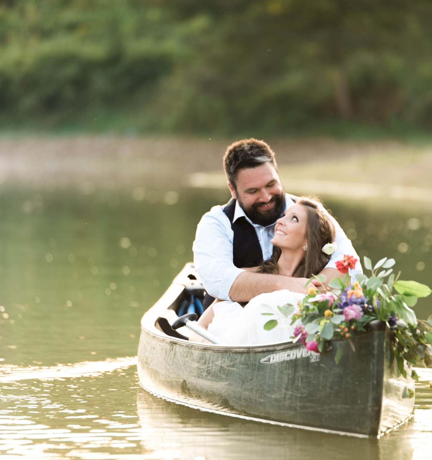 A couple is sitting in a canoe on calm water, surrounded by greenery, with the woman holding a bouquet.