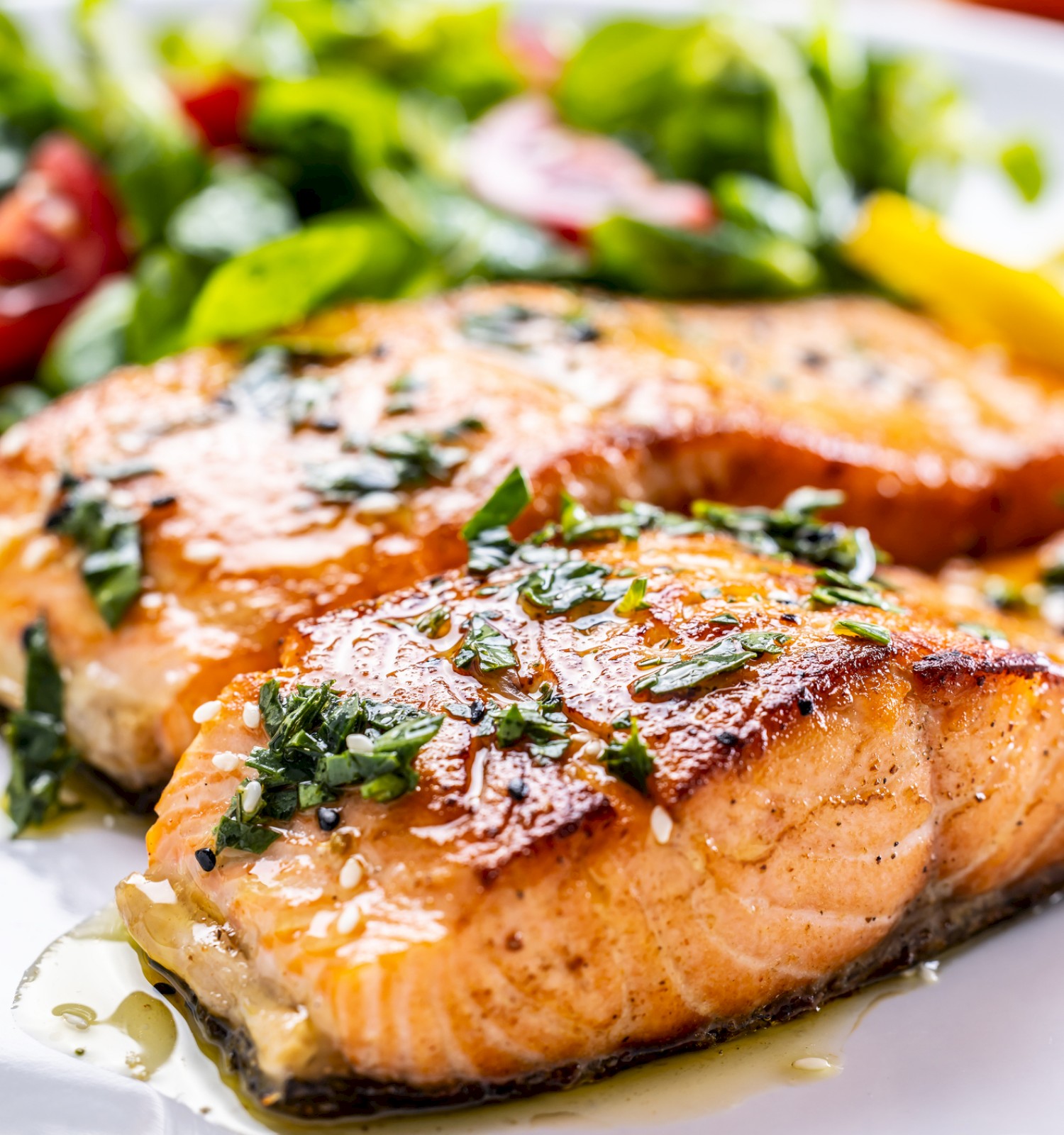 Grilled salmon fillets seasoned with herbs, served alongside a fresh mixed green salad on a white plate.