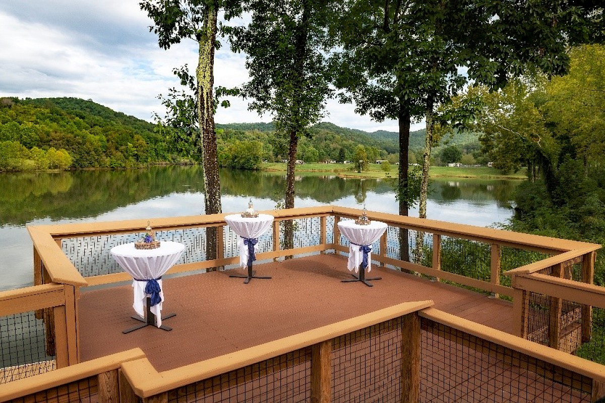 A wooden deck overlooking a serene lake with tall trees, featuring three small tables with white tablecloths arranged on the deck.