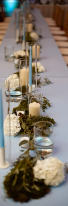 A long table with blue cloth, decorated with candles, leaves, white flowers, and water-filled glasses, surrounded by wooden chairs.