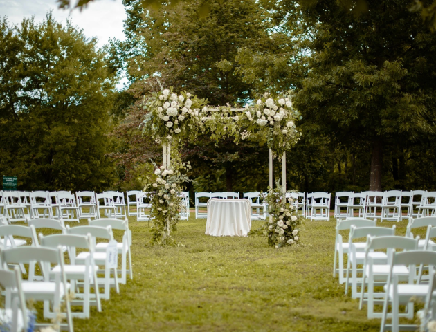 An outdoor wedding setup with white chairs, a floral arch, and a table, surrounded by trees.