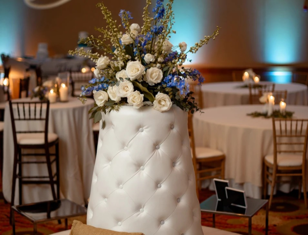 A round seating area with a tufted white centerpiece, topped with white and blue flowers, and a decorative pillow in an elegant setting.