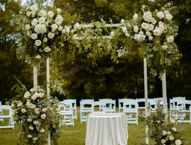 A floral wedding arch adorned with white flowers and greenery stands on a grassy area, with white chairs arranged behind it.