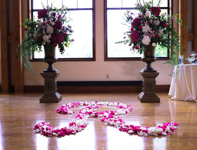 Large floral arrangements flank windows on wooden stands, with a heart made of rose petals on the floor. A small table is nearby.