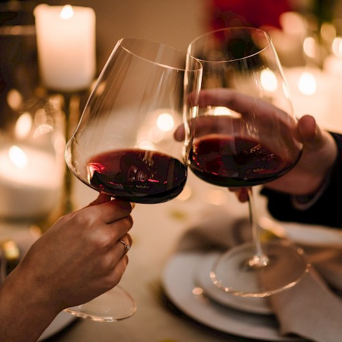 Two people enjoying a candlelit dinner, clinking glasses of red wine with a romantic ambiance in the background.