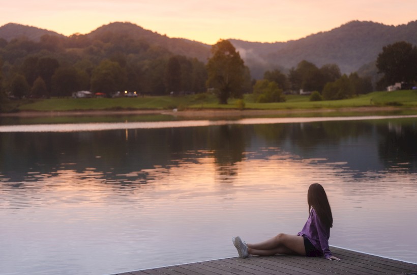 A girl in a purple jacket sits on a dock by a calm lake at sunset, surrounded by mountains and greenery in a peaceful outdoor scene.