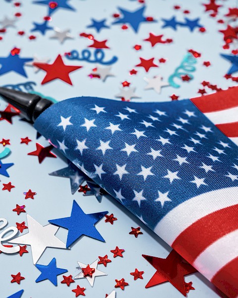 An American flag among red, white, and blue star-shaped confetti and curly decorations on a light background.