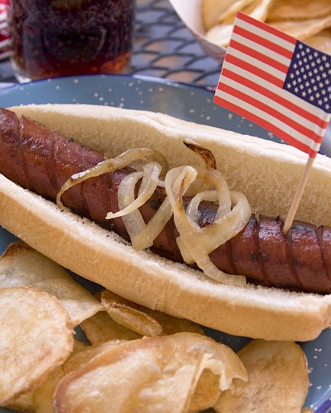A grilled hot dog in a bun topped with onions and a small American flag, served with potato chips on a plate.