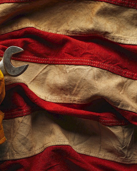 A gloved hand holding wrenches emerges from a wrinkled American flag, symbolizing labor and patriotism.