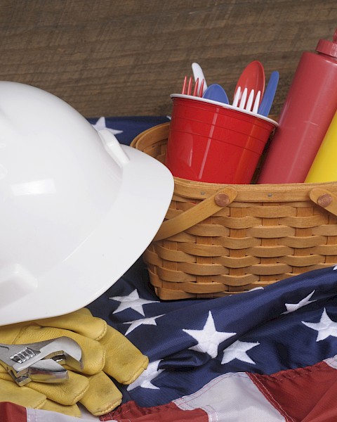 A white hard hat, wrench, gloves, basket with condiments, and utensils on an American flag-themed cloth.
