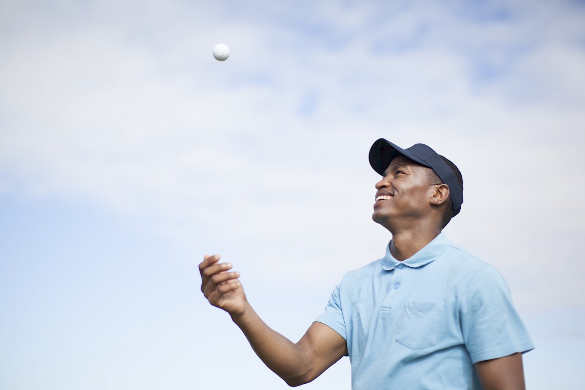A person in a light blue shirt and cap is tossing a ball into the air, looking up at it against a blue sky.