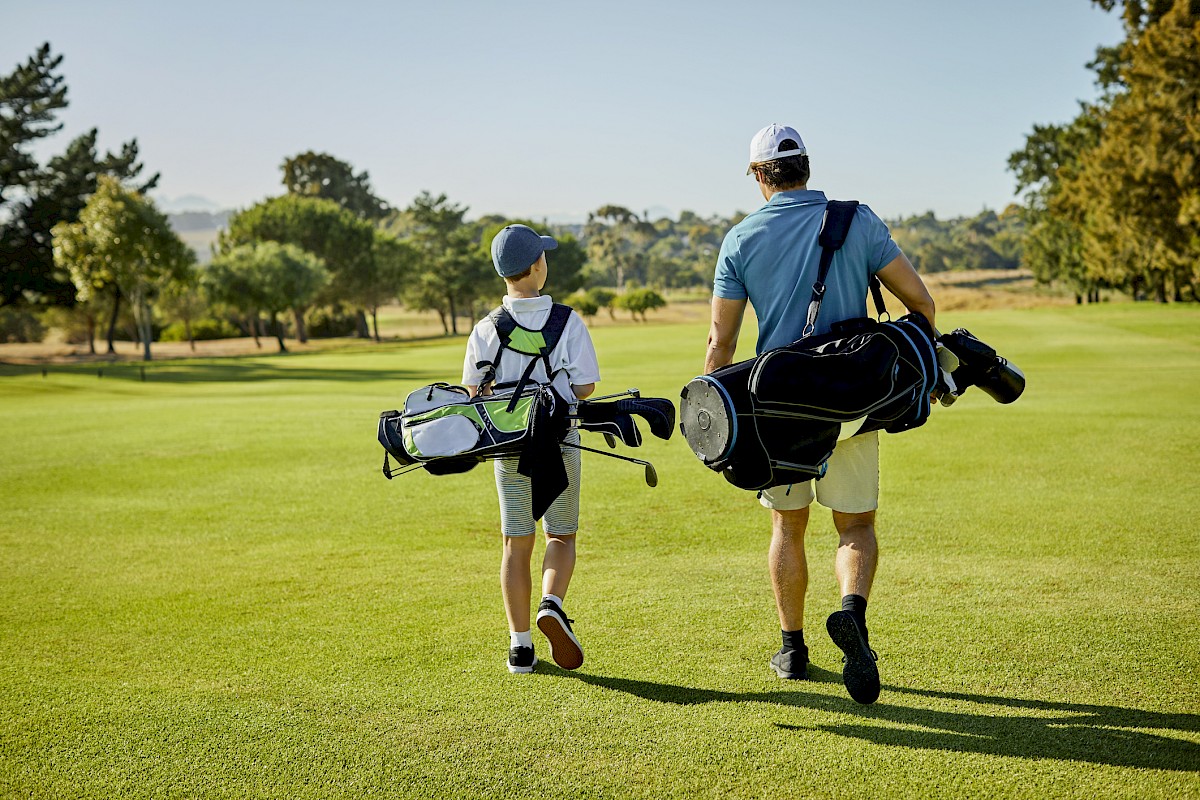 Two people carrying golf bags walk across a lush green golf course, surrounded by trees, under a clear sky.