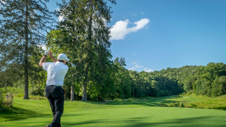 A golfer in white prepares to swing on a lush, green golf course under a bright blue sky with trees and hills in the background.