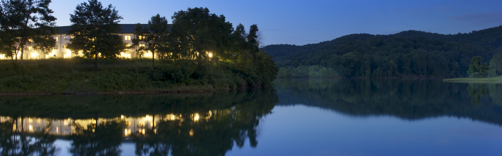 A serene lake at dusk with a building and trees reflected in the water, under a clear, deep blue sky.