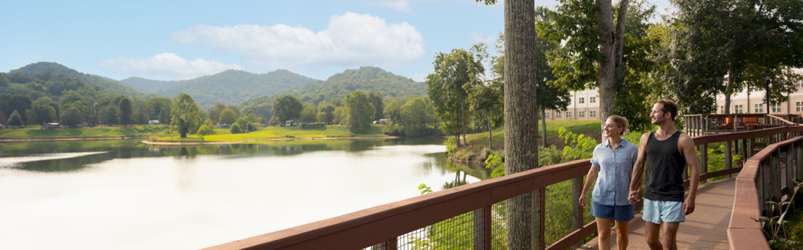 A couple walks hand in hand on a wooden trail beside a tranquil lake with mountains and a blue sky in the background.