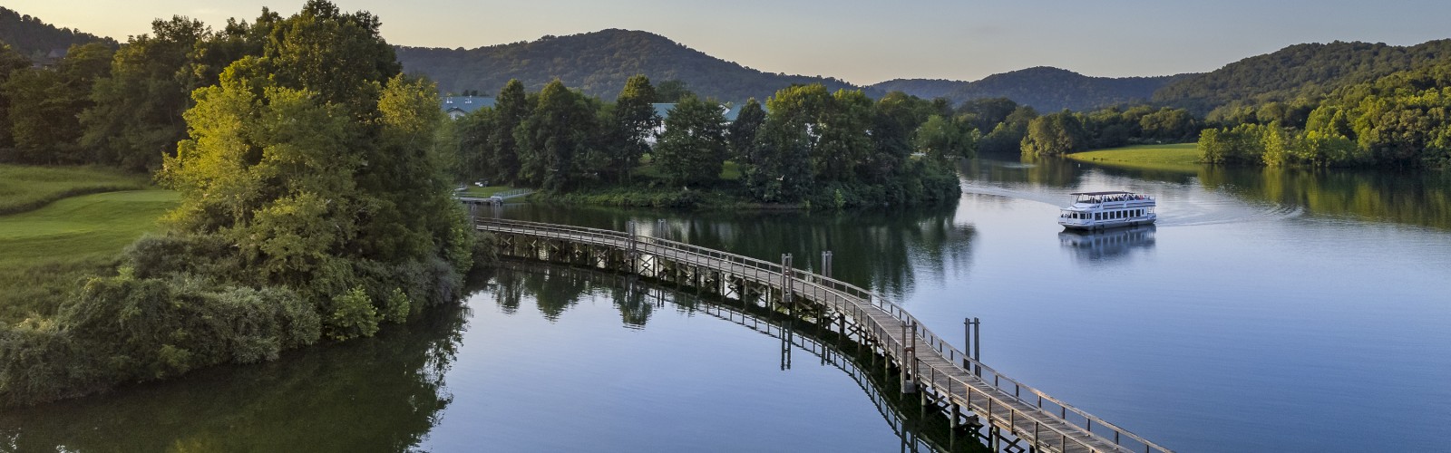 A serene lakeside view with a winding boardwalk, trees, hills, and a small boat on the tranquil water under a clear sky.