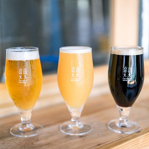 Three glasses of beer in varying shades from light to dark on a wooden surface, with a blurred background.