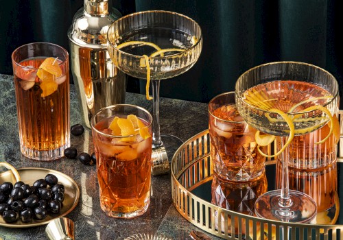An elegant cocktail setup with various drinks garnished with citrus peels, a shaker, olives, and a gold tray on a dark surface.