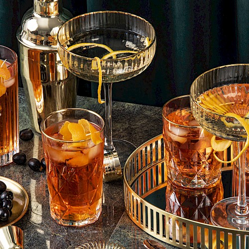 An elegant cocktail setup with various drinks garnished with citrus peels, a shaker, olives, and a gold tray on a dark surface.