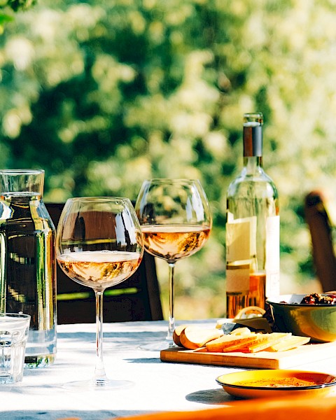 A table set outdoors with wine glasses, a bottle, water carafe, and snacks like cheese, olives, and bread on a sunny day.