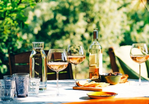 A table set outdoors with wine glasses, a bottle, water carafe, and snacks like cheese, olives, and bread on a sunny day.