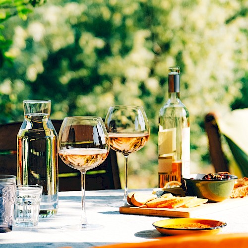 A table set outdoors with wine glasses, a bottle, water carafe, and snacks like cheese, olives, and bread on a sunny day.