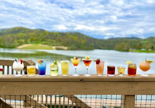 An assortment of colorful cocktails lined up on a wooden railing, with a scenic lake and greenery in the background.