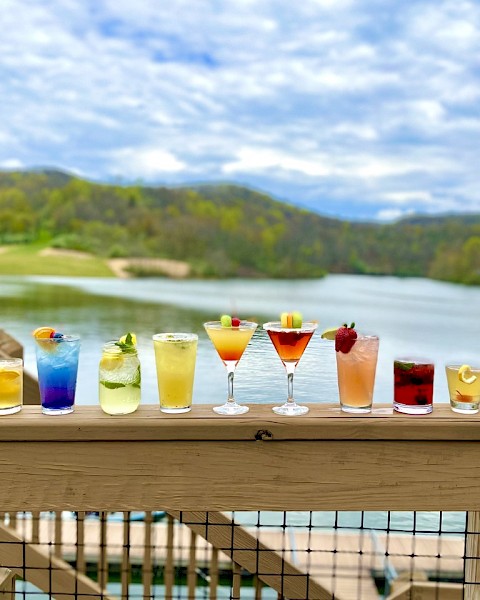 A row of colorful cocktails on a wooden deck overlooking a scenic lake and hills under a partly cloudy sky.