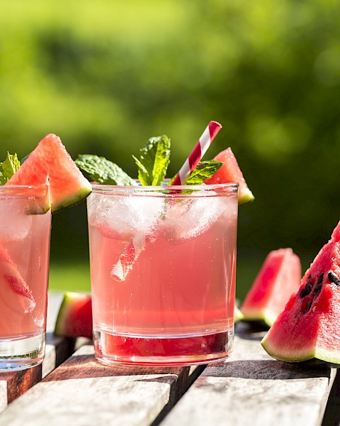 Two glasses of watermelon drinks with ice, garnished with watermelon slices and mint, placed on a wooden table outside.