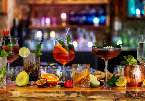 An array of colorful cocktails with garnishes like lime, mint, and berries on a bar counter, surrounded by sliced fruits and herbs.
